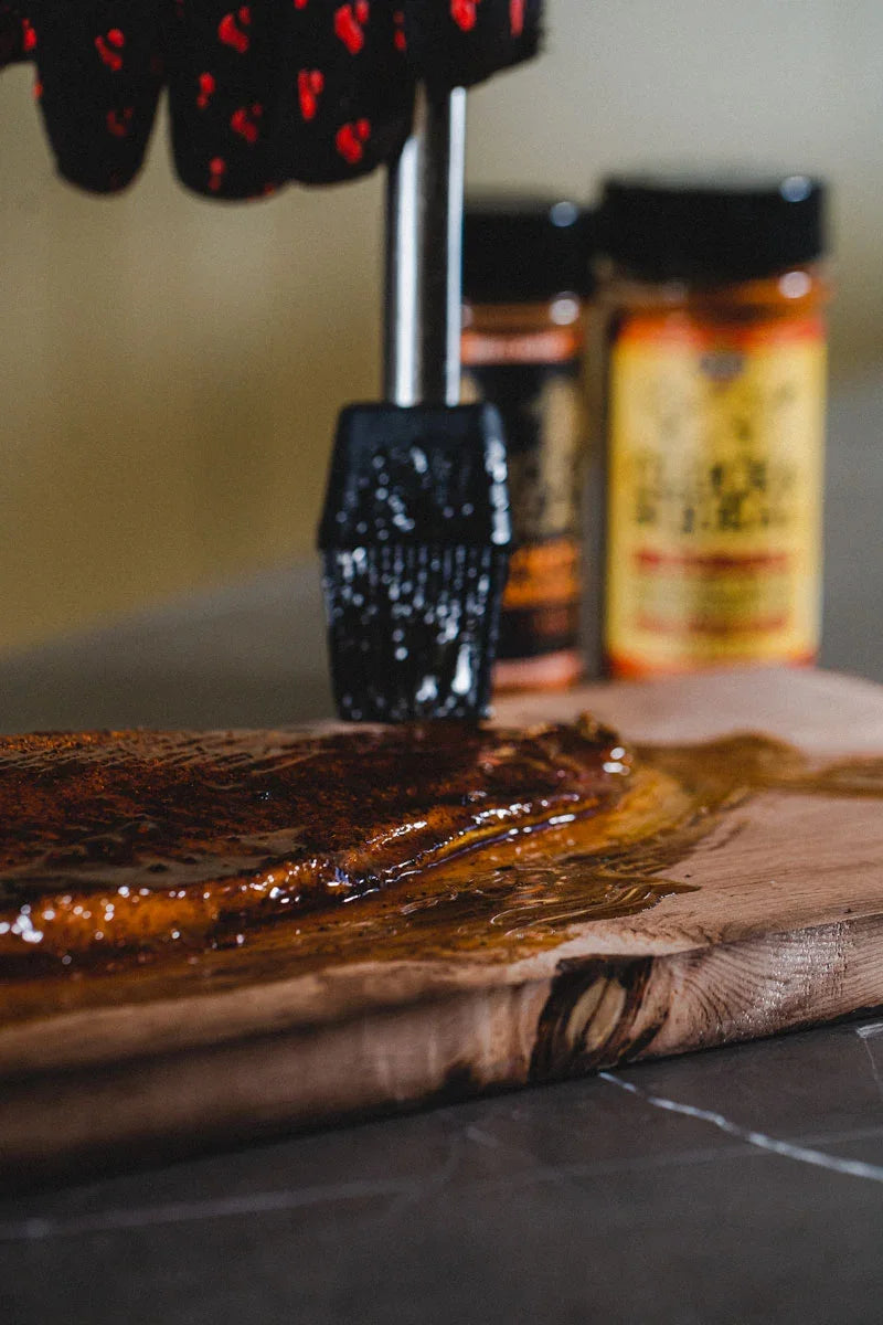 BBQ rub being brushed onto seasoned meat with all-natural seasoning jars in background